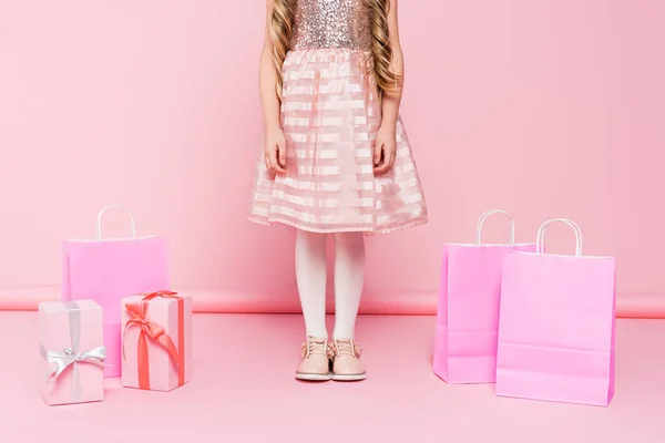 Vista recortada de la niña en vestido de pie cerca de regalos y bolsas de compras en rosa - foto de stock