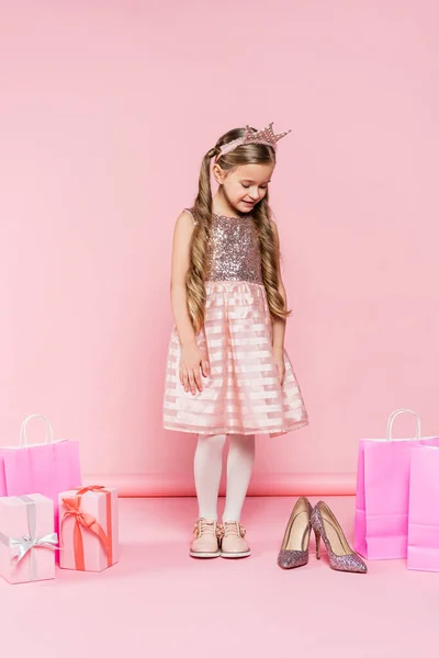 Pleine longueur de petite fille heureuse en couronne regardant les talons près des cadeaux et des sacs à provisions sur rose — Photo de stock