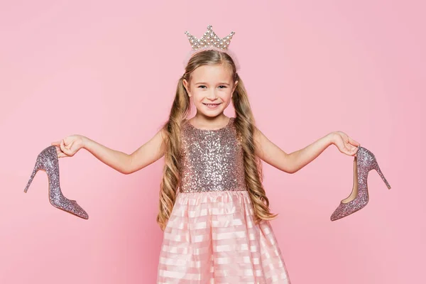 Веселая маленькая девочка в короне и платье держа каблуки изолированы на розовый — стоковое фото