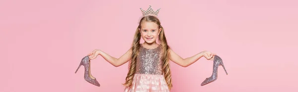Весела маленька дівчинка в короні і сукні тримає підбори ізольовані на рожевому, банер — стокове фото