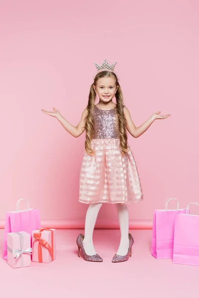 In voller Länge glückliches kleines Mädchen in Krone, das auf Absätzen neben Geschenken und Einkaufstaschen auf rosa steht — Stockfoto