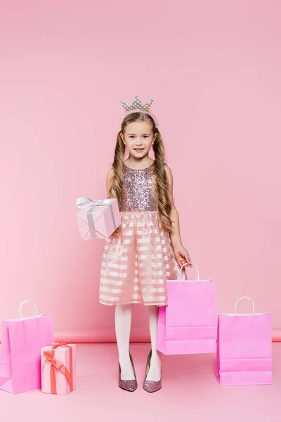 Full length of happy little girl in crown standing on heels and holding present and shopping bag on pink — Stock Photo
