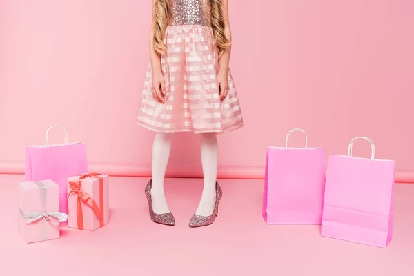Vue recadrée de la petite fille en robe debout sur les talons près des cadeaux et des sacs à provisions sur rose — Photo de stock
