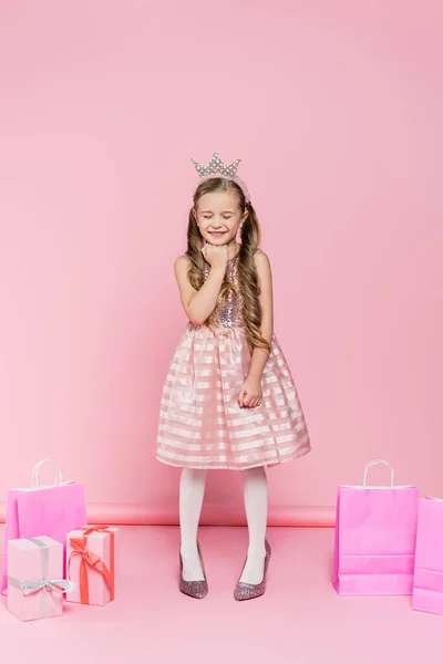 In voller Länge glückliches kleines Mädchen in Krone, das auf Absätzen neben Geschenken und Einkaufstaschen auf rosa steht — Stockfoto