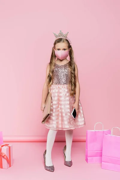 Full length of little girl in crown and medical mask standing on heels, holding smartphone and bag near present and shopping bags on pink — Stock Photo