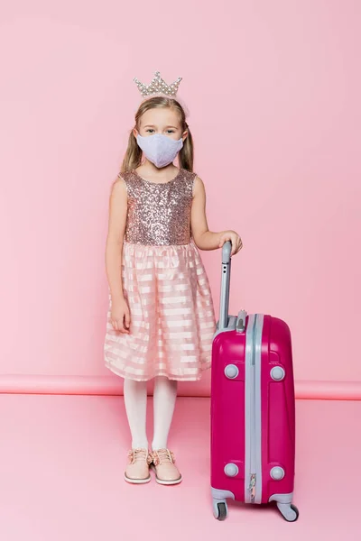 Full length of little girl in crown and medical mask standing near baggage on pink — Stock Photo
