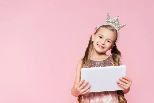 Glückliches kleines Mädchen in Krone hält digitale Tablette isoliert auf rosa — Stockfoto