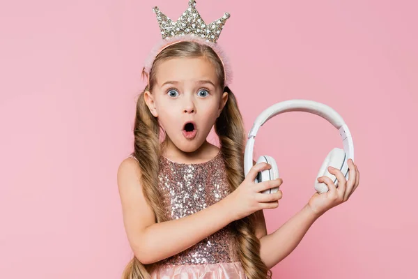 Shocked little girl in crown holding wireless headphones isolated on pink — Stock Photo