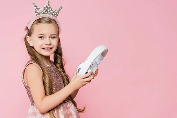 Счастливая маленькая девочка в короне держа беспроводные наушники изолированы на розовый — стоковое фото