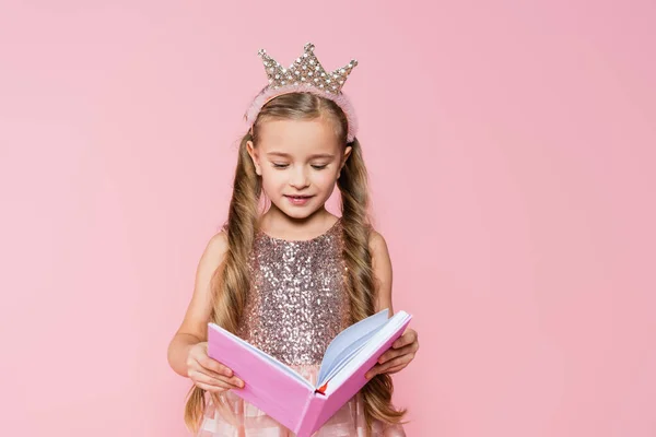 Happy little girl in crown reading book isolated on pink — Stock Photo