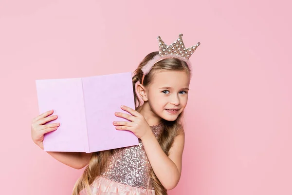 Menina feliz em coroa segurando livro isolado em rosa — Fotografia de Stock