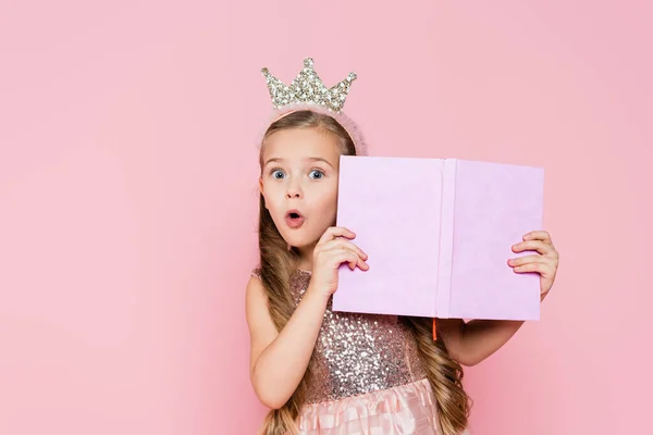 Sorprendida niña en corona sosteniendo libro aislado en rosa - foto de stock