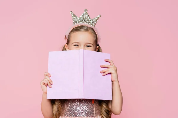 Niña en la cara cubierta de corona con libro aislado en rosa - foto de stock