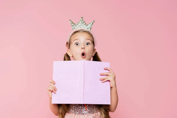 Impactado niña en corona celebración libro mientras mira cámara aislada en rosa - foto de stock