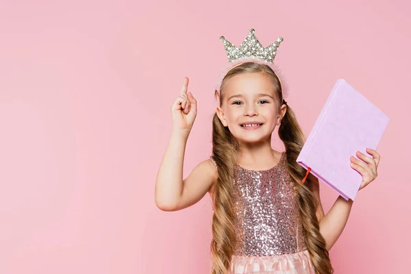 Sonriente niña en corona sosteniendo libro y señalando con el dedo aislado en rosa - foto de stock