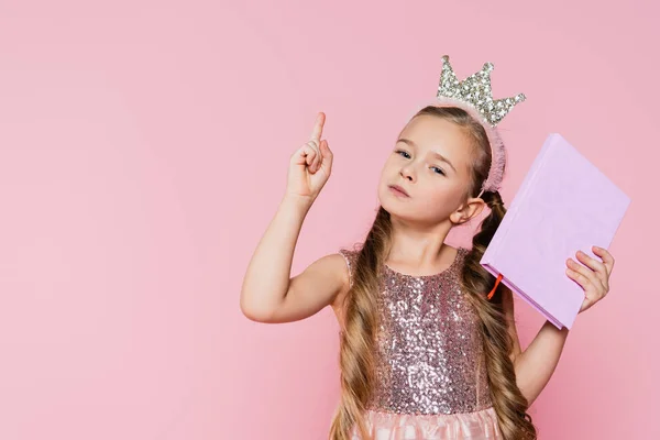 Niña en corona sosteniendo el libro y señalando con el dedo aislado en rosa - foto de stock