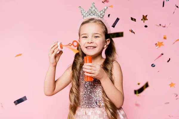 Happy little girl in crown holding soap bubbles near falling confetti on pink — Stock Photo