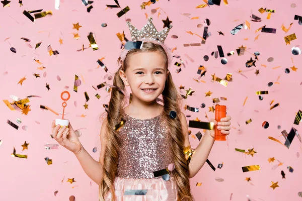 Allegra bambina in corona che tiene bolle di sapone vicino a cadere coriandoli su rosa — Foto stock