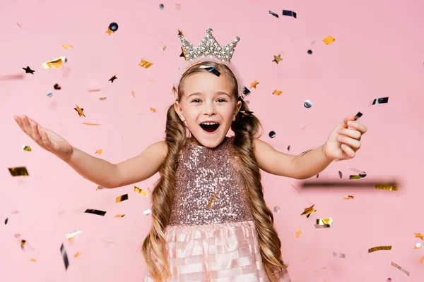 Petite fille étonnante en couronne avec les mains tendues près de confettis tombant sur rose — Photo de stock