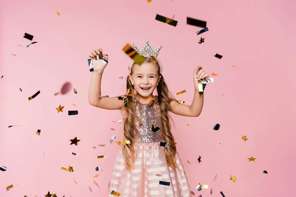 Cheerful little girl in crown and dress near falling confetti on pink — Stock Photo