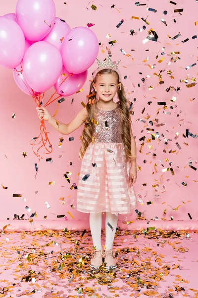 In voller Länge glückliches kleines Mädchen in Krone mit Luftballons in der Nähe fallenden Konfetti auf rosa — Stockfoto