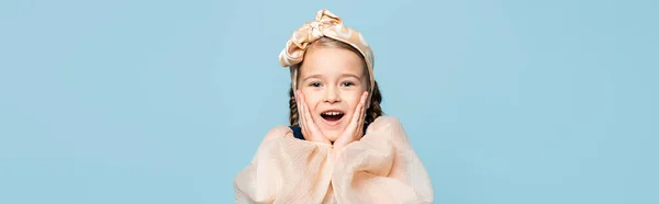 Amazed kid in headband with bow touching cheeks isolated on blue, banner — Stock Photo