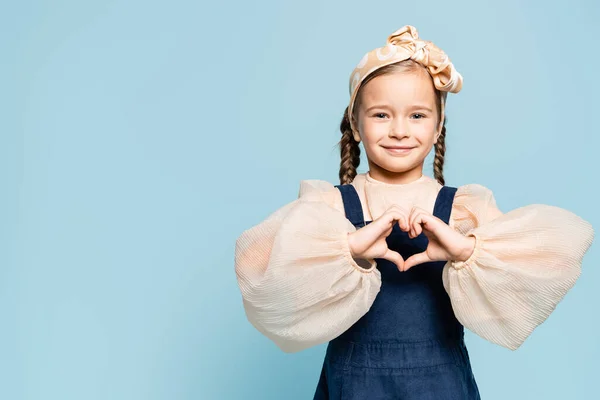 Fröhliches Kind im Stirnband mit Schleife, das in die Kamera blickt und Herz mit auf Blau isolierten Händen zeigt — Stockfoto