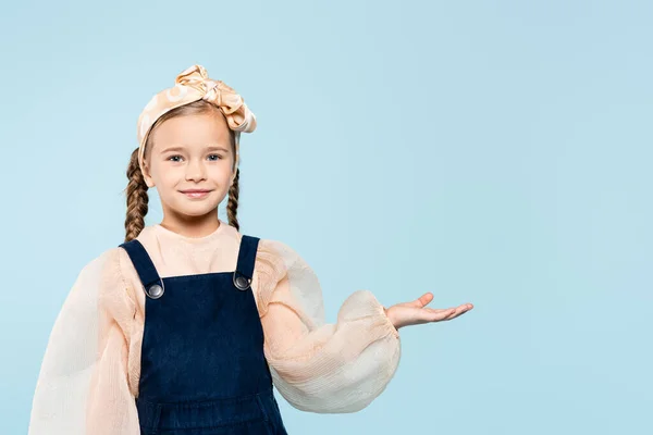 Fröhliches Kind im Stirnband mit Schleife, das in die Kamera schaut, während es mit der Hand isoliert auf blau zeigt — Stockfoto