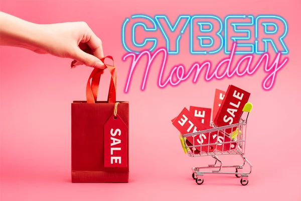 Ausgeschnittene Ansicht einer Frau mit kleiner Einkaufstasche in der Nähe des Warenkorbs mit Verkaufsetiketten in der Nähe von Cyber-Monday-Schriftzug auf rosa — Stockfoto
