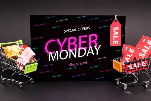 Small presents in toy shopping carts near placard with special offers, cyber monday, shop now lettering on dark background — Stock Photo