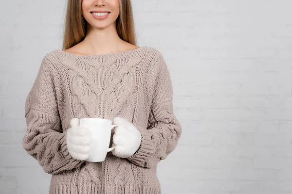 Vista cortada de mulher sorridente em camisola de malha e luvas segurando copo no fundo branco — Fotografia de Stock