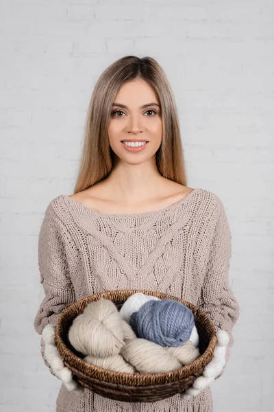 Mujer joven en suéter y guantes sonriendo mientras sostiene la cesta con hilos de punto sobre fondo blanco — Stock Photo