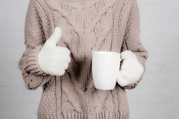 Обрізаний вид молодої жінки в в'язаному светрі і теплих рукавичках, що тримають чашку і показують великий палець на білому тлі — стокове фото