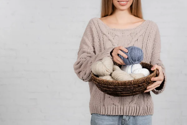 Cropped view of smiling woman in sweater holding yarn and basket on white background — Stock Photo