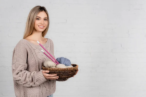 Mulher sorridente segurando cesta com agulhas de tricô e bolas de fios de lã no fundo branco — Fotografia de Stock