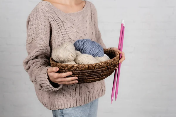 Vista ritagliata della donna in maglione che tiene il cesto con fili di lana e ferri da maglia su sfondo bianco — Foto stock