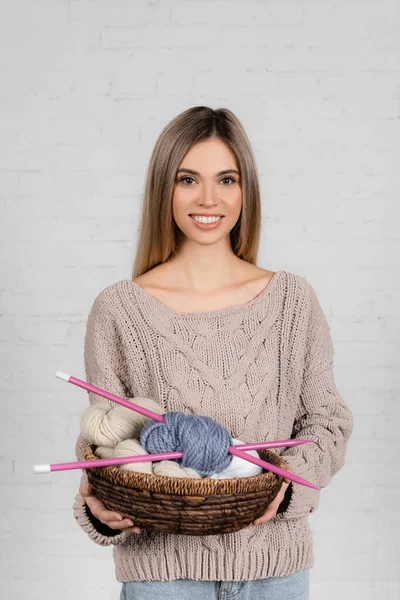 Smiling woman holding basket with woolen yarn and needles with brick wall at background — Stock Photo