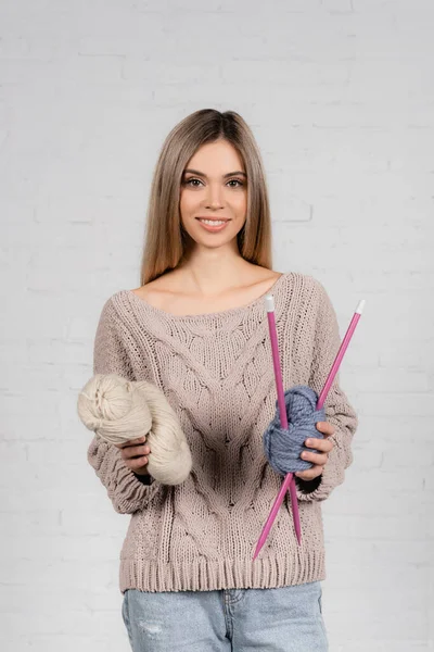 Mujer sonriente en suéter sosteniendo hilo de lana y agujas de punto sobre fondo blanco - foto de stock