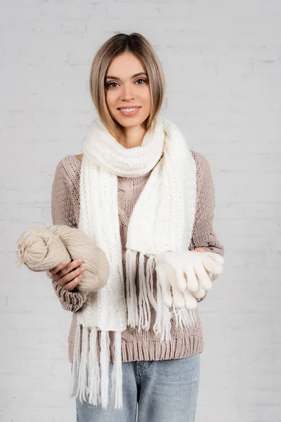 Femme souriante en pull et écharpe tenant des gants chauds et du fil sur fond blanc — Photo de stock