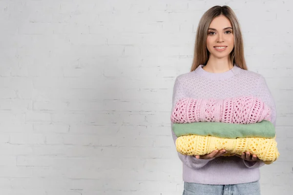 Young woman smiling at camera while holding colorful sweaters with white brick wall at background — Stock Photo