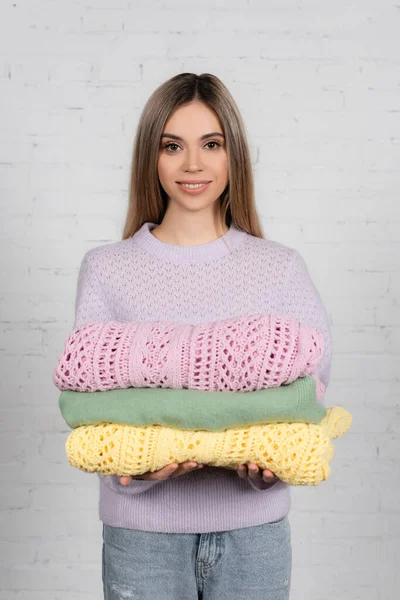 Smiling woman holding warm sweaters and looking at camera near white brick wall — Stock Photo