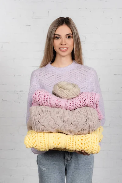 Mujer sonriente mirando a la cámara mientras sostiene suéteres calientes e hilo sobre fondo blanco - foto de stock