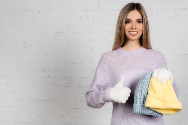 Donna sorridente in maglione lavorato a maglia e guanti caldi che reggono cappelli e mostrano come vicino al muro di mattoni bianchi — Foto stock