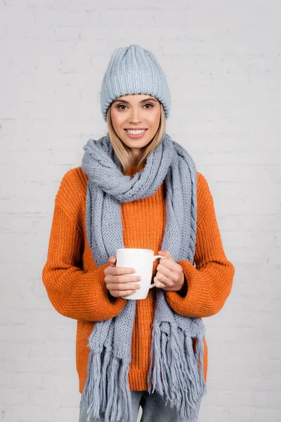 Mujer joven en jersey de punto, sombrero y bufanda sosteniendo la taza y sonriendo sobre fondo blanco - foto de stock