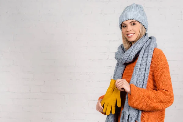 Cheerful woman in knitted wear holding gloves near white brick wall — Stock Photo