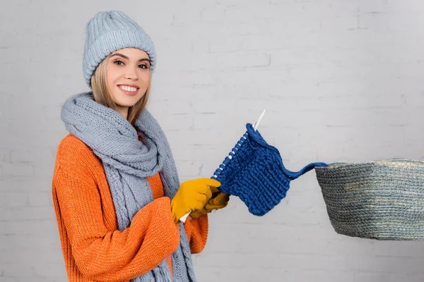 Femme joyeuse regardant la caméra tout en tricotant près du panier et mur de briques blanches sur fond blanc — Photo de stock