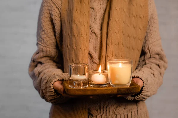 Cropped view of burning candles on wooden board in hands of woman in scarf and sweater blurred on white background — Stock Photo