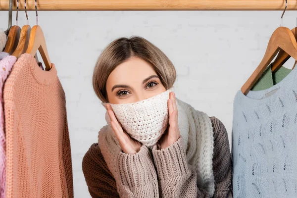 Жінка вкриває обличчя шарфом біля светрів на вішалці на білому тлі — стокове фото