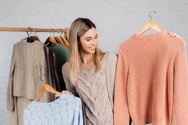 Smiling woman holding hangers with sweaters near hanger rack blurred on white background — Stock Photo