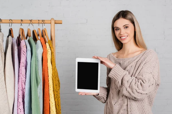 Smiling woman holding digital tablet with blank screen near hanger rack with sweaters on white background — Stock Photo